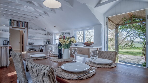 Une salle à manger spacieuse, avec une table en bois dressée pour quatre personnes, entourée de chaises en osier. On aperçoit, au fond, une cuisine avec des meubles blancs et une grande fenêtre.