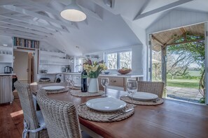 Uma ampla sala de jantar com uma mesa de madeira posta para quatro pessoas, rodeada por cadeiras de vime. Uma cozinha com armários brancos e uma janela grande é visível ao fundo.
