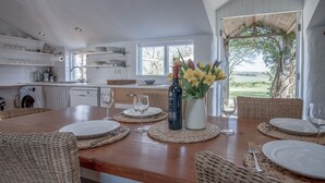 Un coin repas avec une table en bois dressée pour quatre personnes, entourée de chaises en osier. Une bouteille de vin et un vase rempli de fleurs sont posés sur la table.