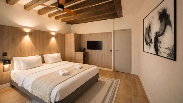 A modern bedroom with a bed, wooden ceiling beams, a ceiling fan, a mounted TV, and a painting on the wall. - Lovely studio for 3 in the heart of the Marais (Paris)