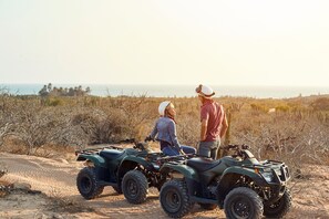 To personer på ATV'er i et tørt, åbent landskab med sparsom vegetation og havudsigt i det fjerne.
