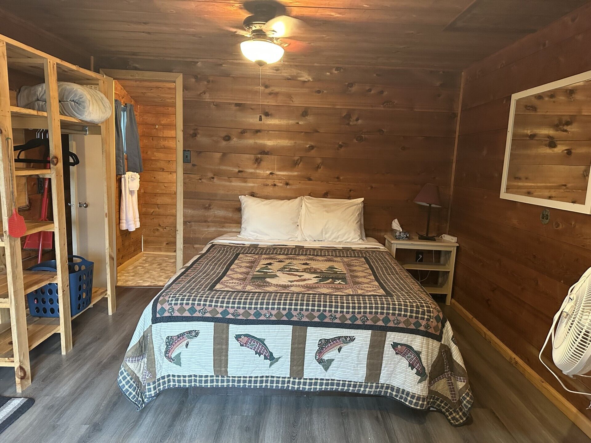 Une chambre confortable avec un plafond en bois, un lit recouvert d’une courtepointe à motifs de poissons, une armoire où sont suspendus des vêtements, et une petite table de chevet avec une lampe.