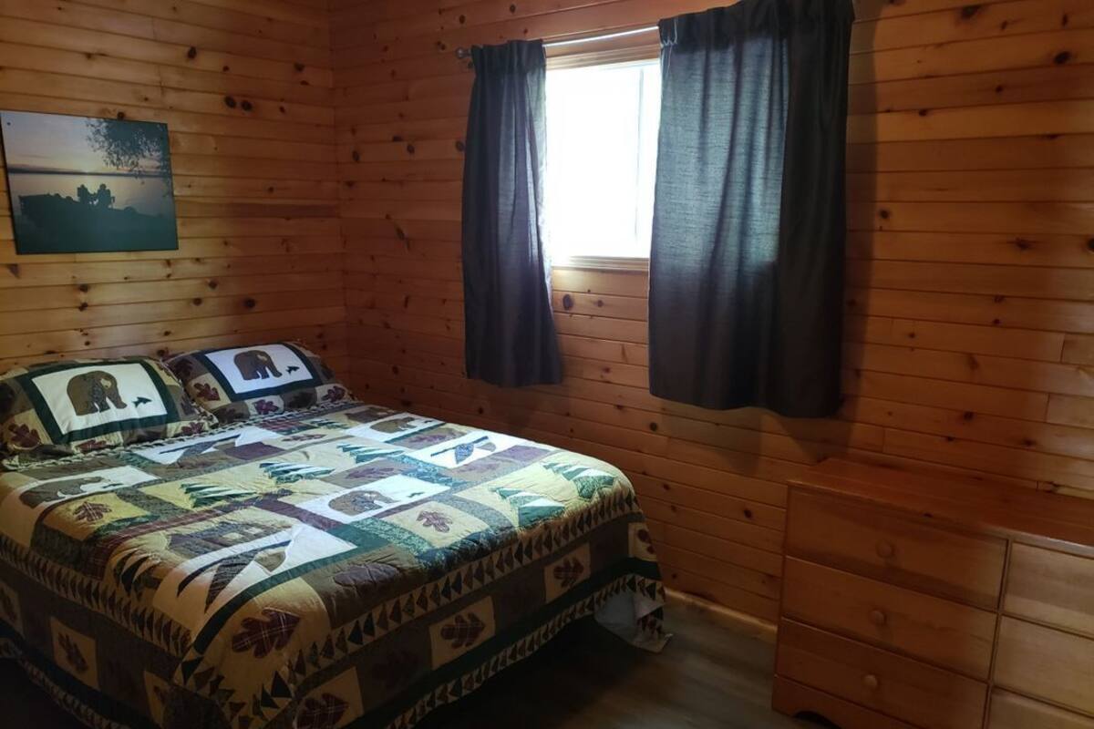 Une chambre dans une cabane en bois, avec une couette matelassée, une table de chevet, une commode et une fenêtre avec des rideaux.
