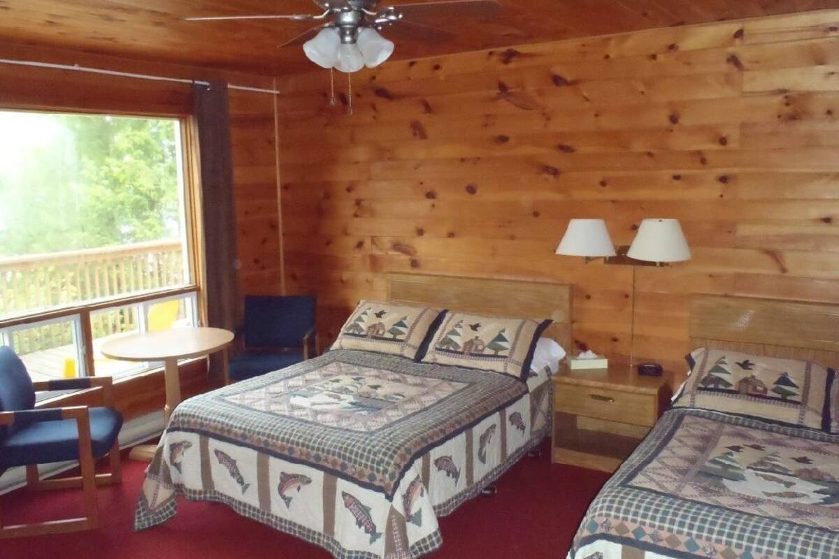 Une chambre dans un chalet en bois, avec deux lits, un ventilateur de plafond, une petite table et une fenêtre donnant sur la forêt.
