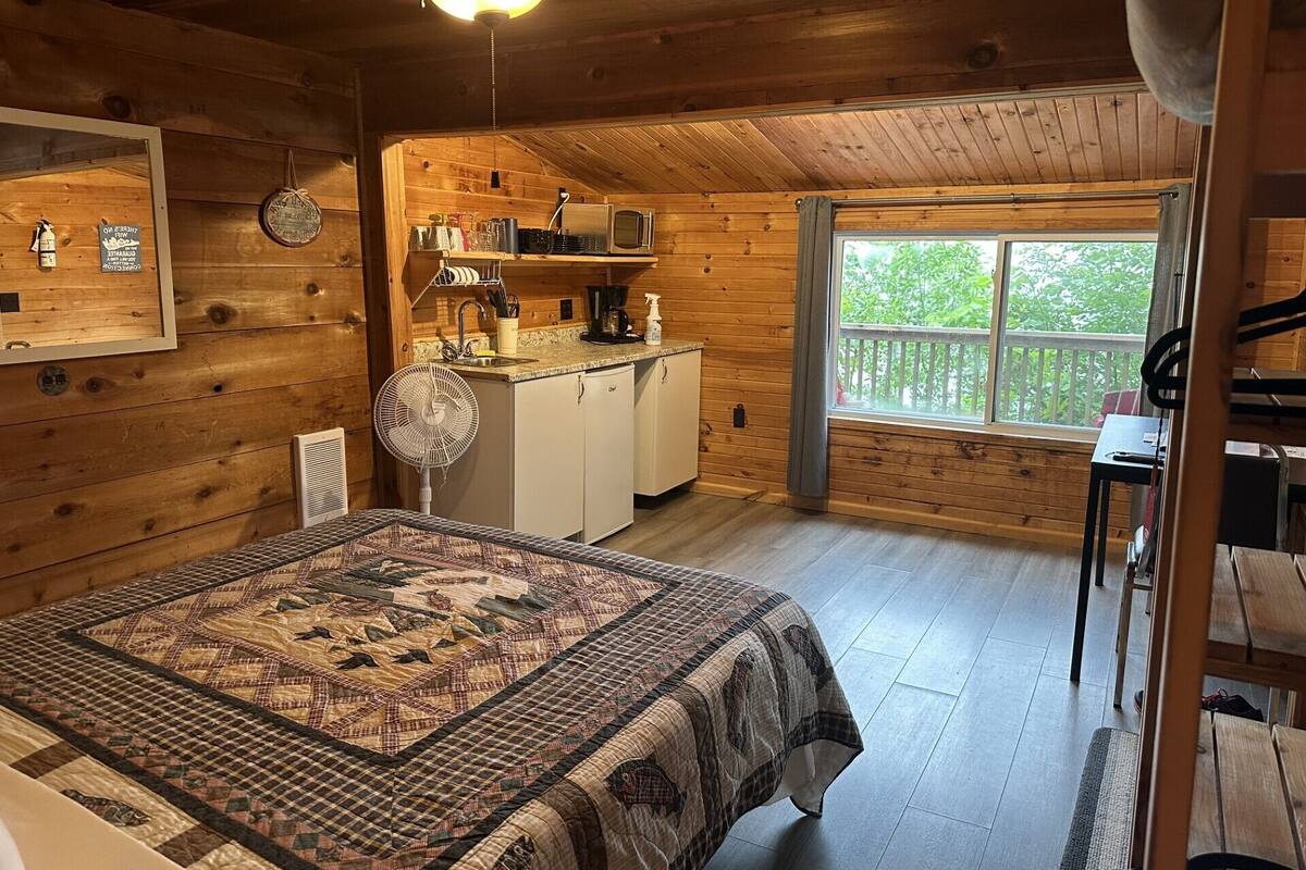 Une chambre confortable, aménagée dans le style d’un chalet, avec un lit, une kitchenette et une grande fenêtre donnant sur un paysage verdoyant.