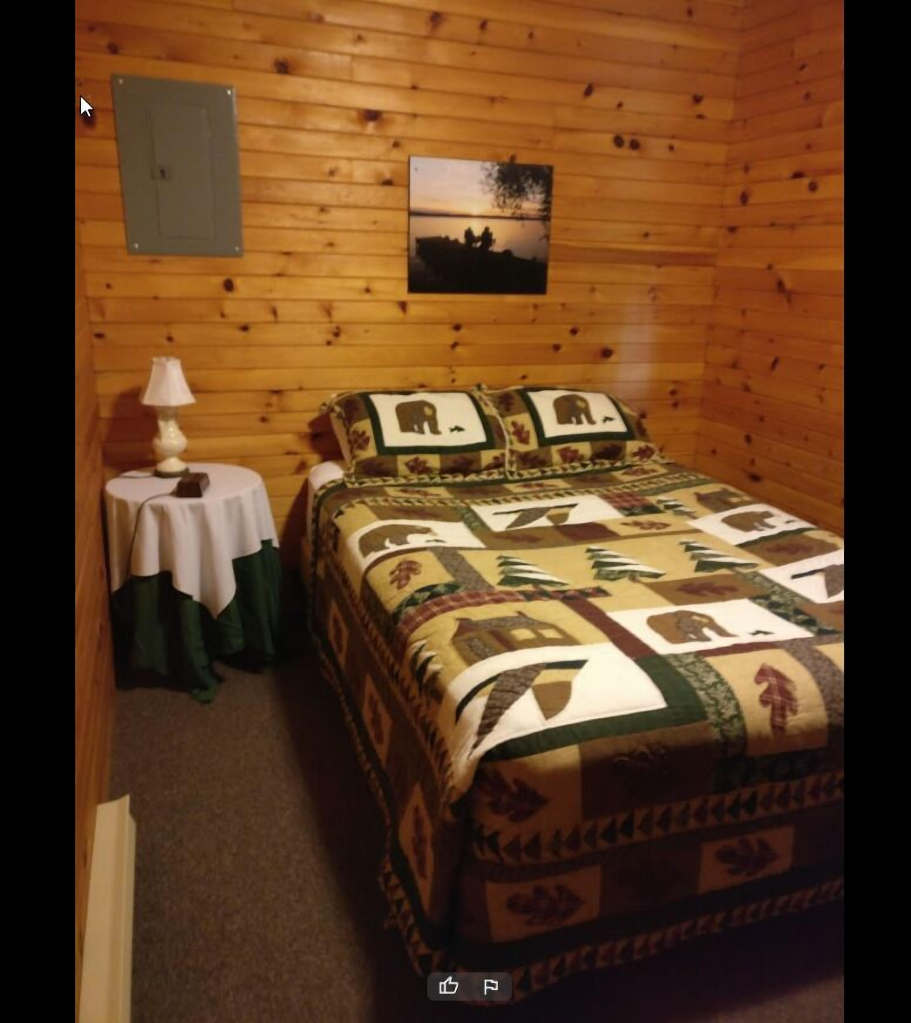 A cozy room with a wooden interior, a bed with a quilt featuring animal prints, a small table with a lamp, and a picture on the wall.