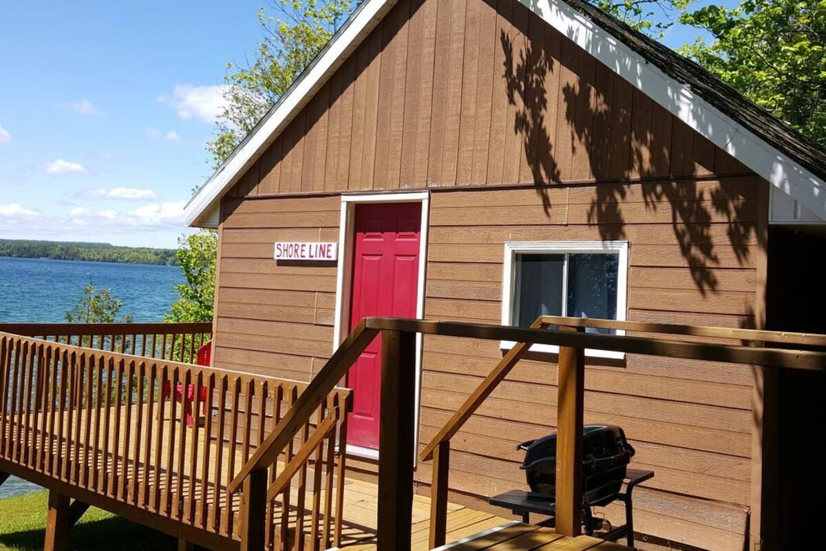 Une cabane en bois avec une porte rouge portant l’inscription « SHORE LINE », une terrasse et un barbecue.