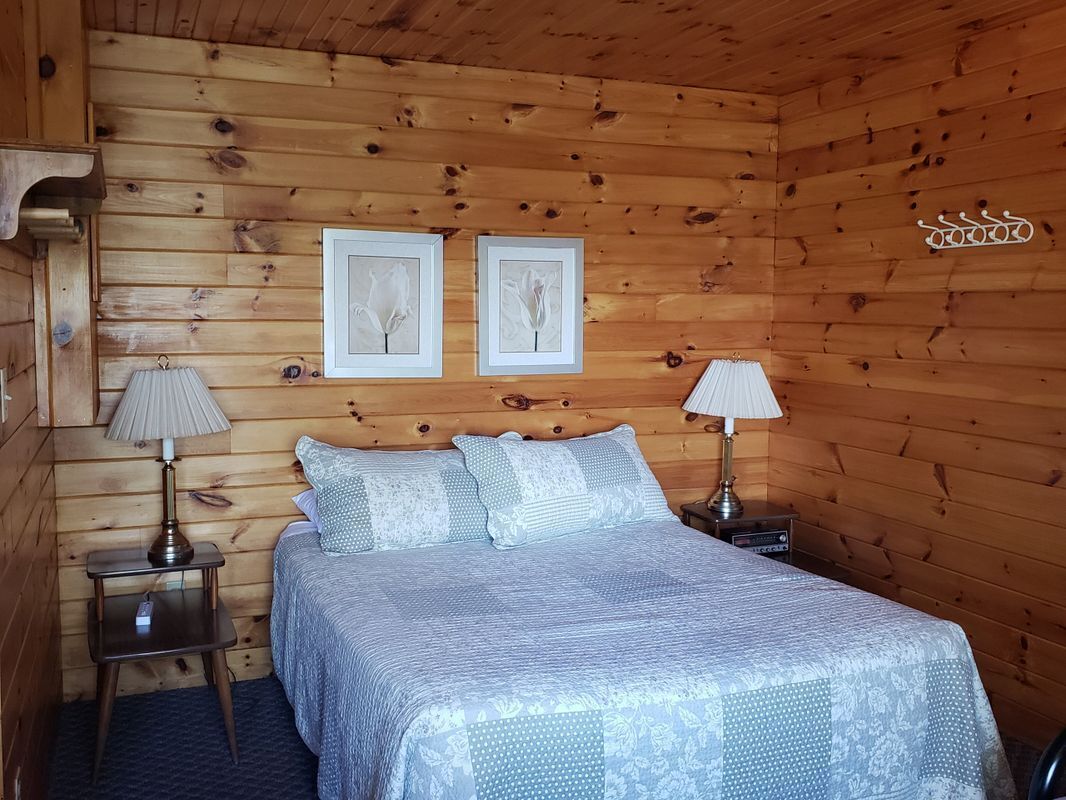 A wooden cabin bedroom with a bed, two bedside lamps, and two framed pictures on the wall.