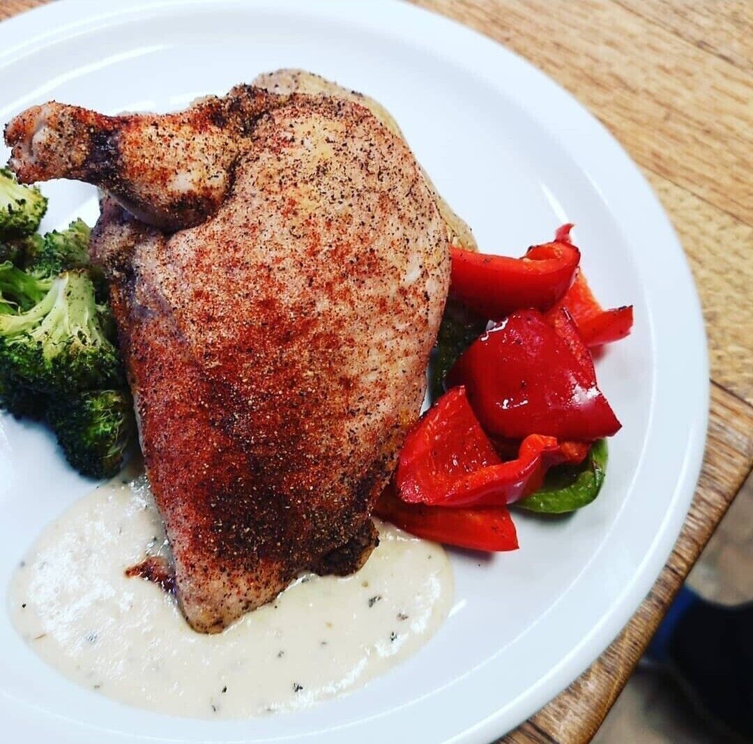 Ein Teller mit einem gebratenen Hähnchenschenkel, Brokkoli, roten Paprika und einer cremigen Soße.