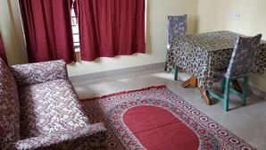 A room with a sofa, a dining table with chairs, a patterned rug, and a window with red curtains. - Serene Peaceful Flat (Kolkata)