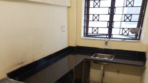 A small kitchen with a black countertop, a window with metal bars, a sink, and a blue trash bin. - Serene Peaceful Flat (Kolkata)
