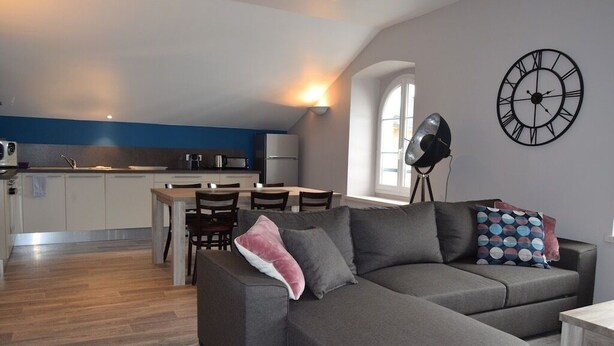 Un salon moderne avec un canapé gris, une table à manger et des chaises. Une grande horloge murale est visible.