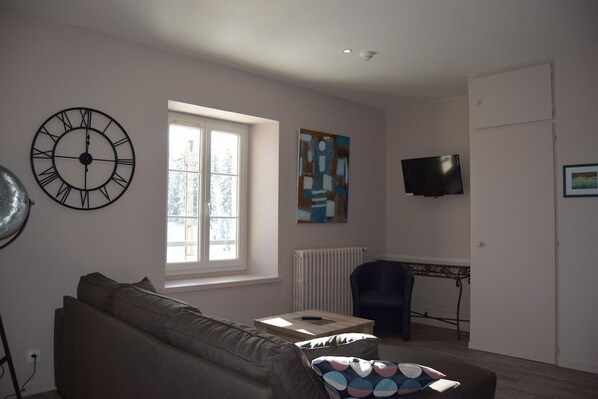 A living room with a sofa, a coffee table, a TV, a large clock, and a window with blinds. - Apartment Coeur du village 5 (Les Rousses)