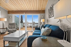 A modern living room with a wooden ceiling, a blue sofa, a white coffee table, and a view through glass doors to an outdoor patio. - Dolphin View-Ocean Front, Multiple pools, Decks overlooking the ocean! (Pine Knoll Shores)