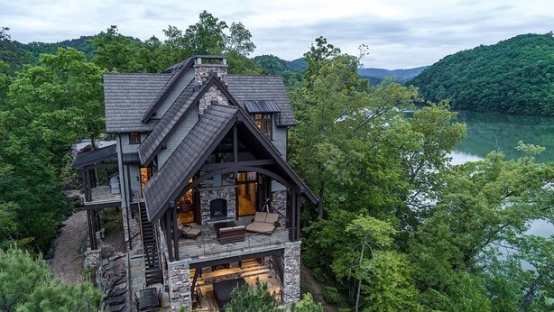 Eine große, mehrstöckige Holzhütte mit Natursteinfassade, Kamin und einer Terrasse mit Blick auf einen See.
