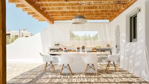 An outdoor dining area with a stone floor, white chairs, and a table set with food and drinks.