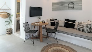 A modern living room with a round wooden table, two black wireframe chairs, a beige sofa with black and patterned cushions, a woven rug, a wall-mounted TV, and a hanging light fixture.