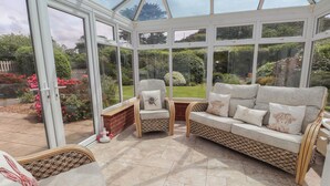A conservatory with a glass ceiling, wicker furniture, and a view of a garden.
