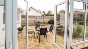 A conservatory with a patio area, including a table and chairs, and a view of a garden and a white house in the background.