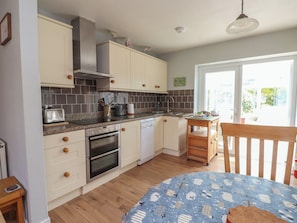 A kitchen with white cabinets, a stove, oven, and a sink. There's a window with a view of greenery outside. - Bro Mebyd (Criccieth)