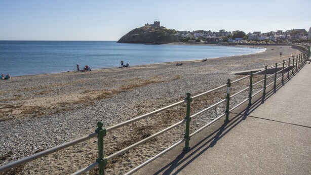 A coastal promenade with a pebbled beach, a green metal railing, and a distant hill with buildings.