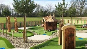 Un campo de minigolf con una cabaña de madera, una estatua y una zona delimitada por una valla.