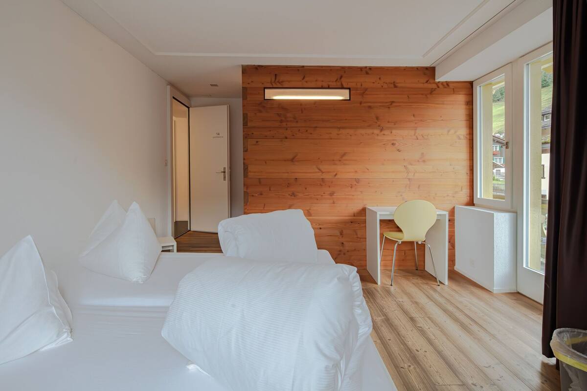 A hotel room with a wooden wall, a bed with white bedding, a desk with a chair, and a window with a view.