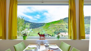 Una mesa de comedor servida con platos, tazas y una cesta de pan, con vistas a un paisaje pintoresco y una figurita de ciervo en el alféizar.