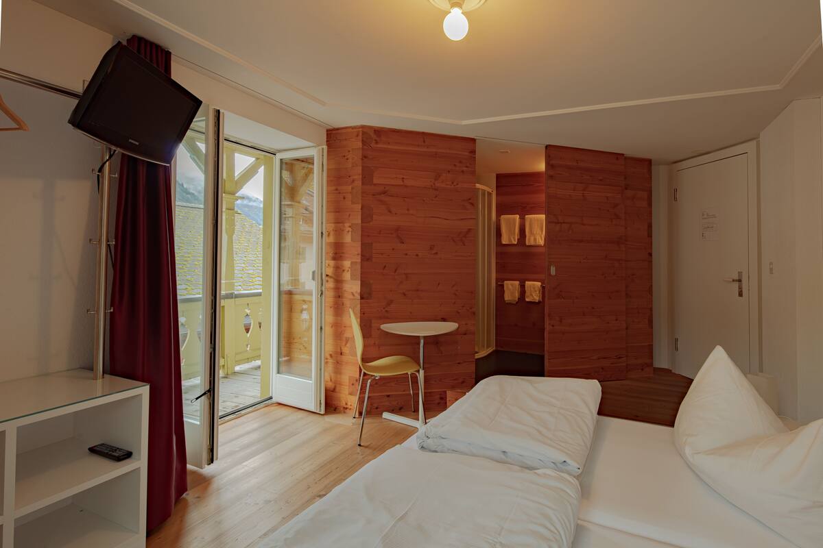 A hotel room with a bed, a small table, a TV mounted on the wall, and a sliding glass door leading to a balcony.