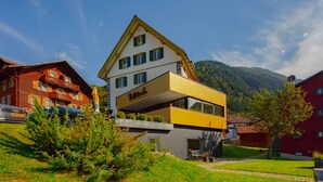 Un hotel con fachada amarilla y blanca, un balcón y montañas al fondo.