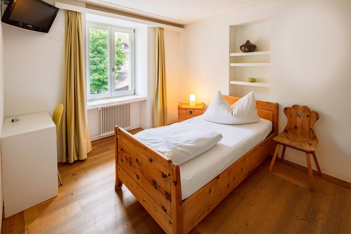 A neatly made bed with a wooden headboard, a bedside table with a lamp, a small wooden chair, and a shelf with decorative items.