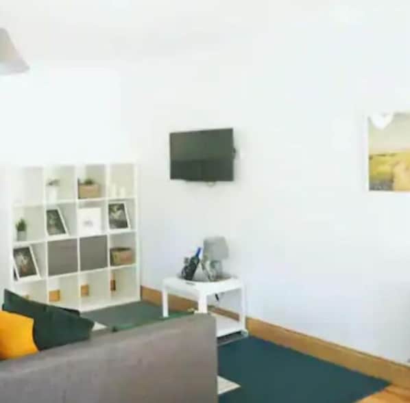 A living room with a grey sofa, a white side table with a lamp, a TV mounted on the wall, and a white shelving unit with various items.