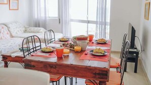 Un comedor con una mesa de madera preparada para el desayuno, incluyendo croissants y jugo de naranja, con un sofá y televisor al fondo.
