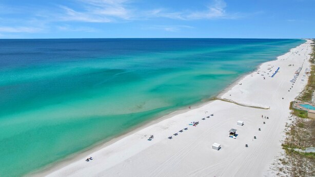 Vue aérienne d’une plage aux eaux turquoise et limpides, au sable blanc, bordée d’une rangée de parasols et de chaises longues.