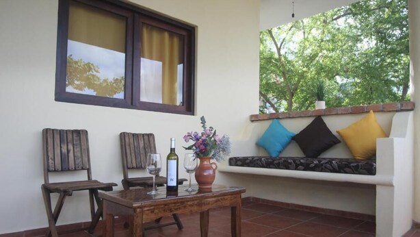 Un patio con una mesa de madera, dos sillas, una botella de vino y un florero con flores. Se ve una ventana con cortinas y vistas a árboles.