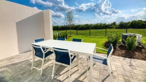 Une terrasse extérieure avec une table blanche et des chaises bleues, entourée d’un mur blanc et offrant une vue sur un champ verdoyant.