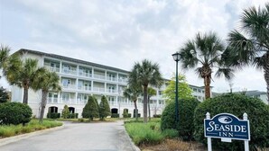 A Seaside Inn building with a white facade, multiple balconies, and a well-maintained garden with palm trees and shrubs.
