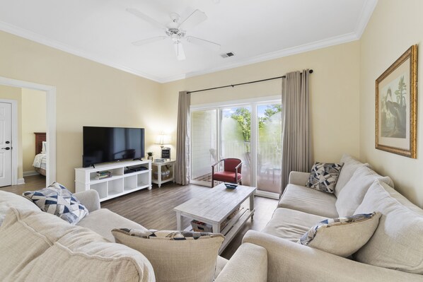 A living room with a flat-screen TV, a white coffee table, beige sofas, a red chair, and a large window with curtains.