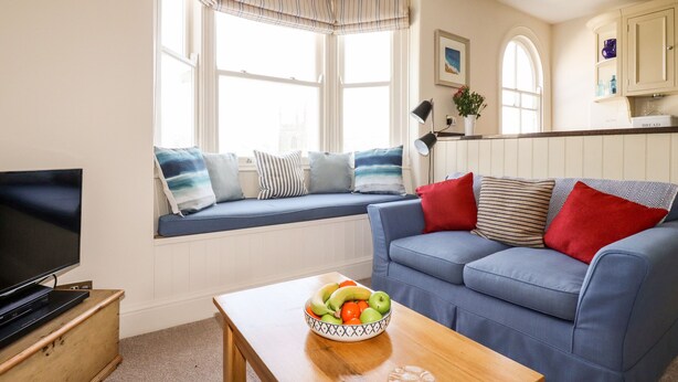 A living room with a sofa, a television, a window seat with cushions, a table with a fruit bowl, and a kitchen area in the background. - Driftwood (Fowey)