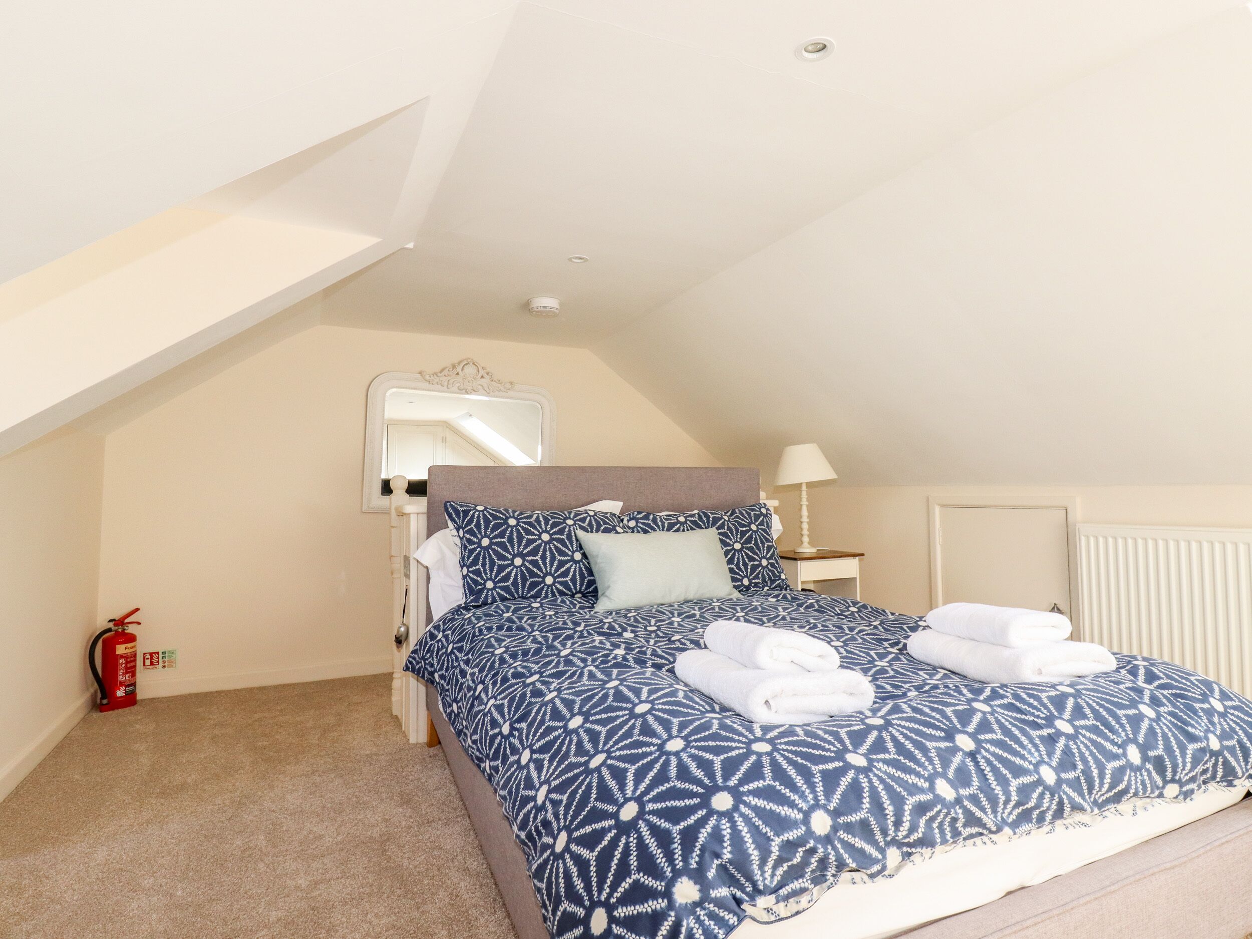 Une chambre avec un plafond en pente, un lit recouvert d’une couvre-lit à motifs, une table de chevet avec une lampe et un extincteur.