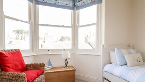A bedroom with a bed, a nightstand, a lamp, a wicker chair, and a view of the street through large windows. - Driftwood (Fowey)