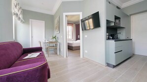 Ein modernes Wohnzimmer mit einem violetten Sofa, ein Essbereich mit Tisch und Stühlen, eine Küche mit Fernseher und Schränken sowie ein durch eine offene Tür einsehbares Schlafzimmer.