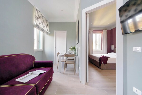 A room with a purple sofa, a dining table with chairs, a bed, and a TV mounted on the wall. - Colosseo Gardens - C13 (Rome)