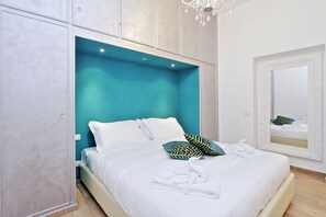 Una cama bien tendida con sábanas blancas y una toalla, una pared de acento color turquesa y un espejo grande con marco decorativo.