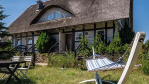 Une maison rustique avec un toit de chaume, une véranda en bois et un hamac dans le jardin.