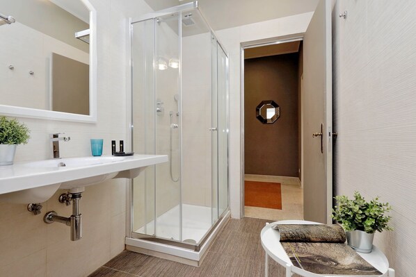 A modern bathroom with a glass shower enclosure, a white sink, and a small seating area with a cushion and a potted plant. - Monti Apartments - M7 (Rome)