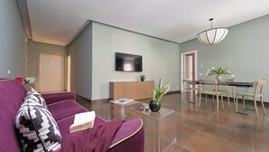 En moderne stue med sofa, sofabord i glas, spiseplads med bord og stole, et fjernsyn ophængt på væggen og en potteplante.