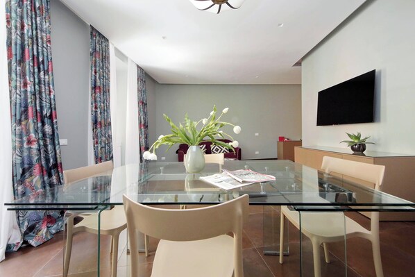 A modern dining area with a glass table, beige chairs, a vase with flowers, and a flat-screen TV mounted on the wall. - Colosseo Gardens - C2 (Rome)