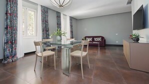 En moderne stue med et glasbord, fire stole, en sofa, et fjernsyn monteret på væggen og et stort vindue med gardiner.