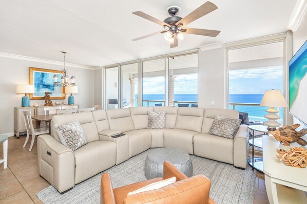 A modern living room with a curved sectional sofa, a dining area with a table and chairs, a ceiling fan, and a large window offering a view of the ocean. - Beautiful 3B Gulf Front w/ Daily Beach Chair Set! (Pensacola Beach)